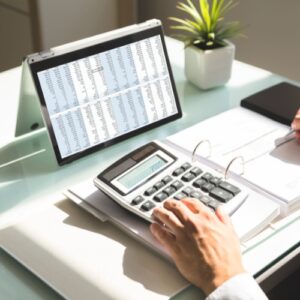 A tablet device displays a spreadsheet with income and expenditure details. On the table rests a calculator and ledger with a person keeping track of the details.