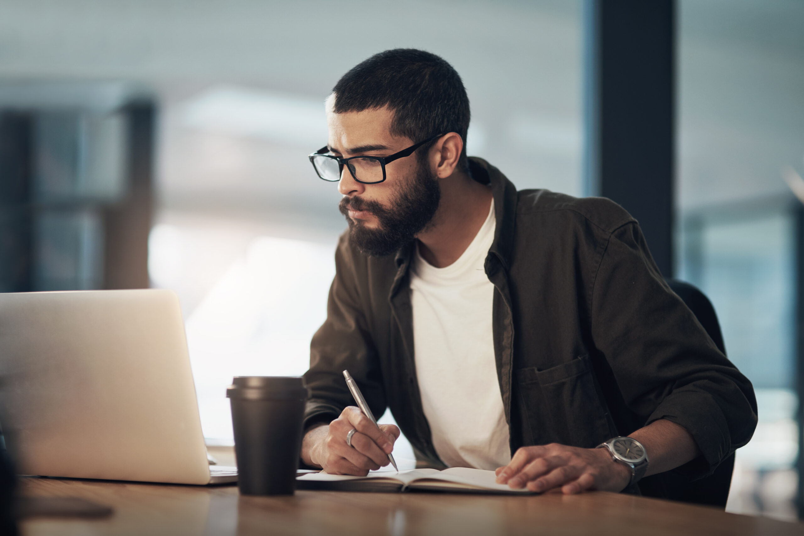Man with glasses and a beard concentrating hard on the screen of his laptop and taking notes