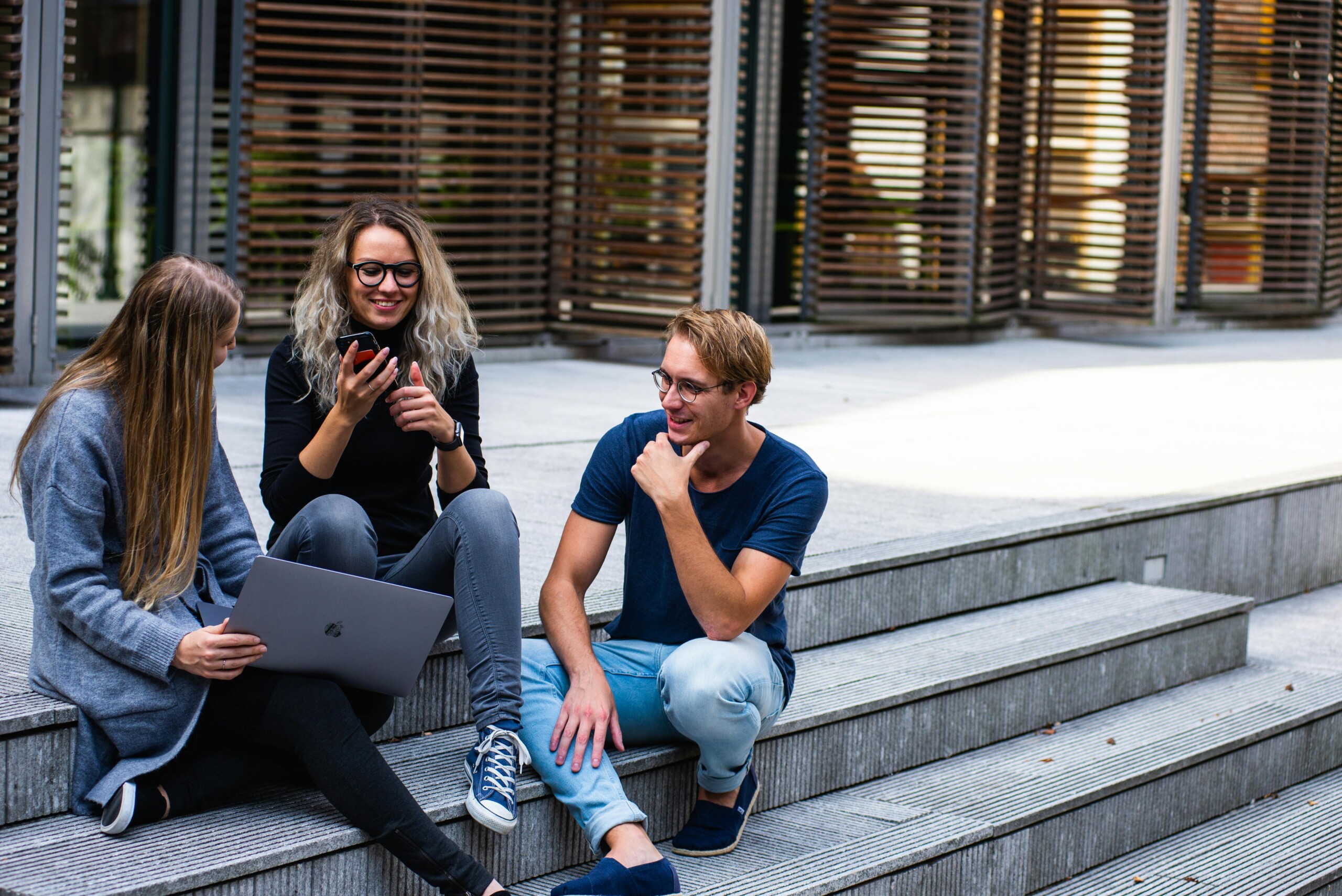 Three people chatting on outdoor steps with a laptop in front of a modern glass building