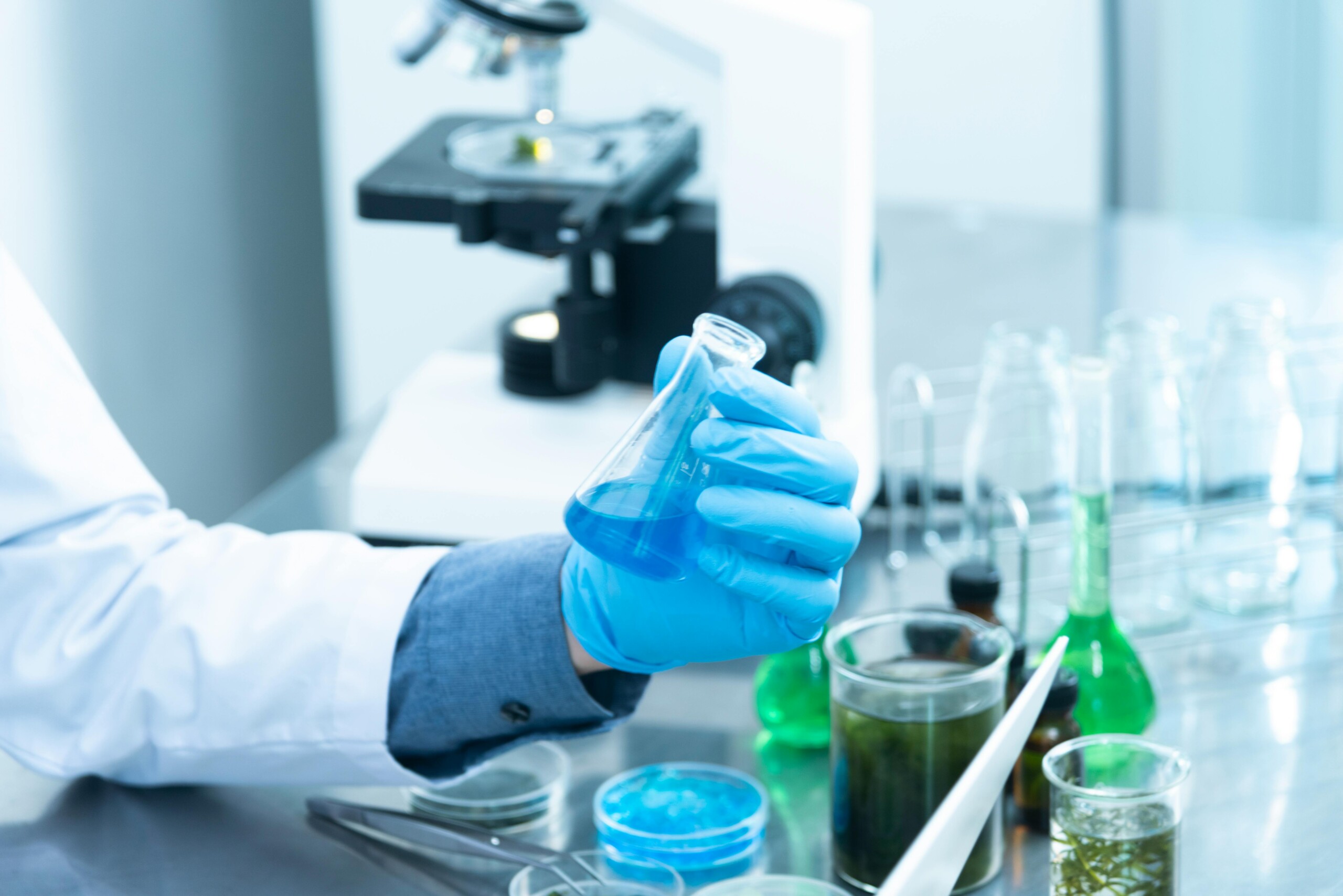 Person in lab coat and gloves holding a flask with blue liquid in a science lab with various chemicals and a microscope.