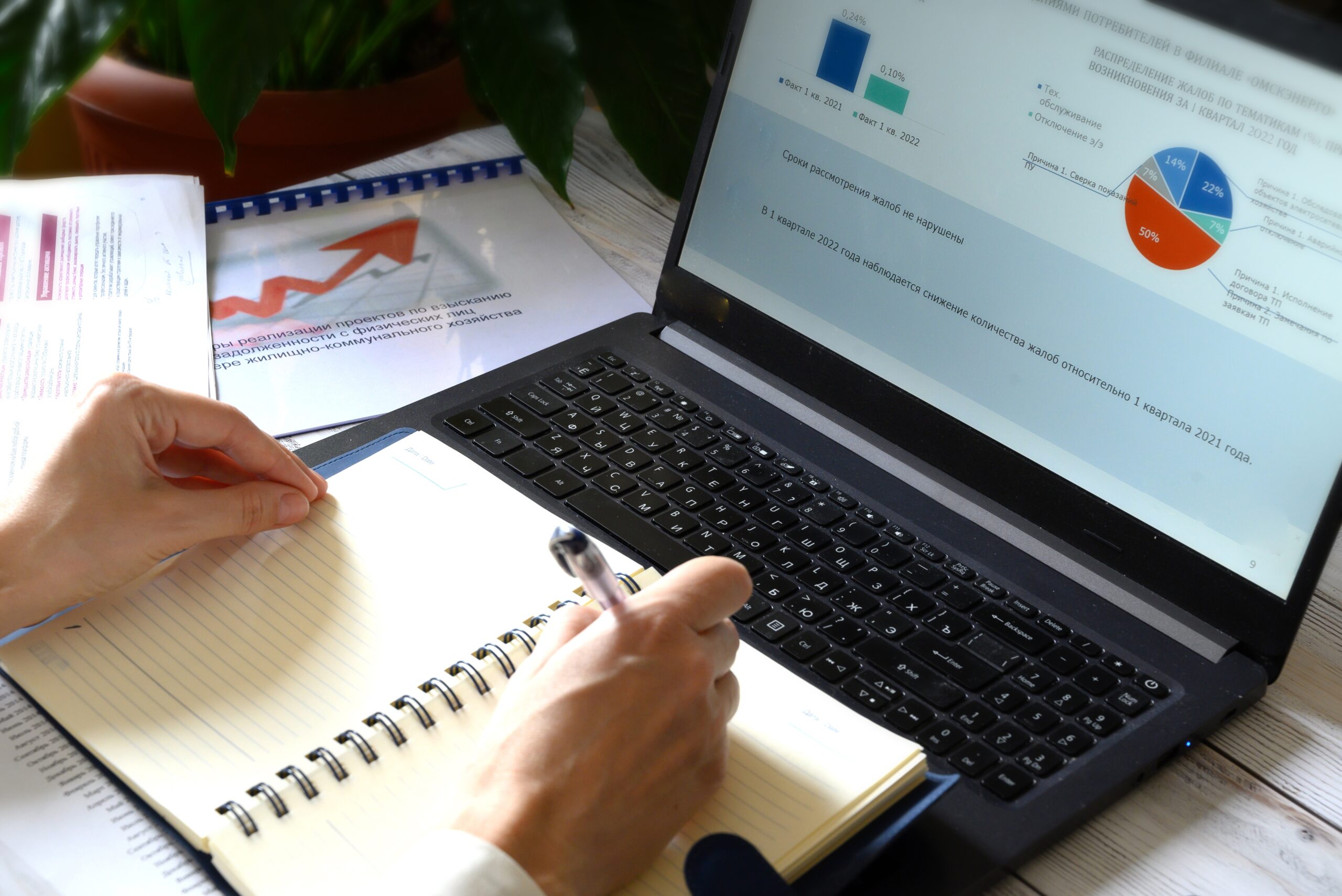 charts displayed on a laptop and a pair of hands writing in a notebook balanced on the keypad