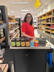 Ruby Chagger standing in selfridges with her jars of curry bases. Ruby wears a red t-shirt and black glasses and is smiling towards the camera in the middle of a retail shopping aisle display showcasing her products.