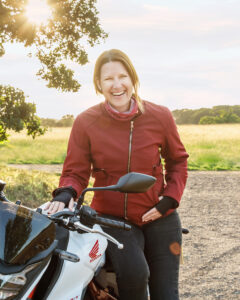 A woman (Linda) smiling and sitting sideways on her motorbike looking at the camera